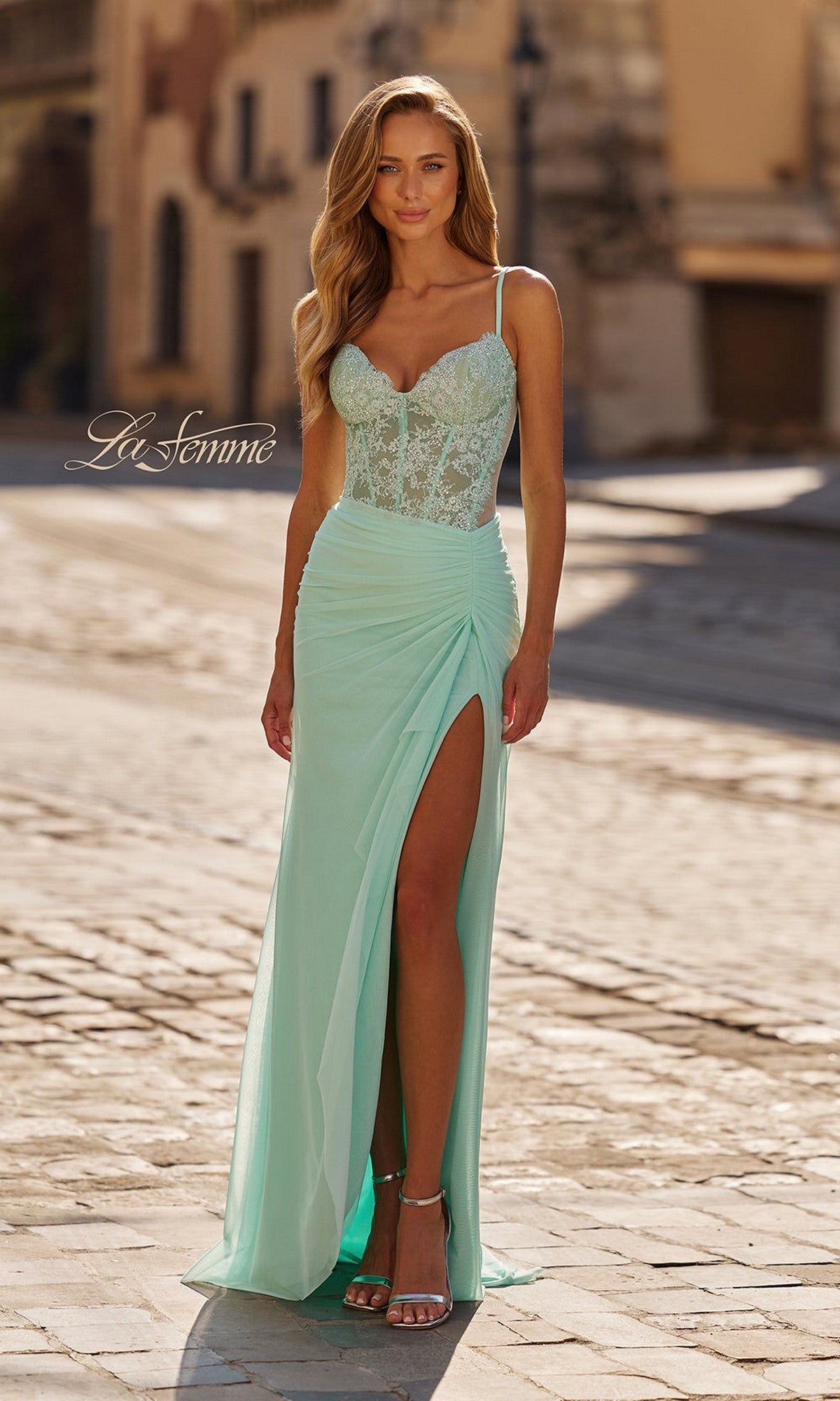 A woman stands on a sunlit cobblestone street in the La Femme 33661 Beaded-Lace-Corset Long Prom Dress, featuring light mint floral beaded lace, spaghetti straps, and a high slit.